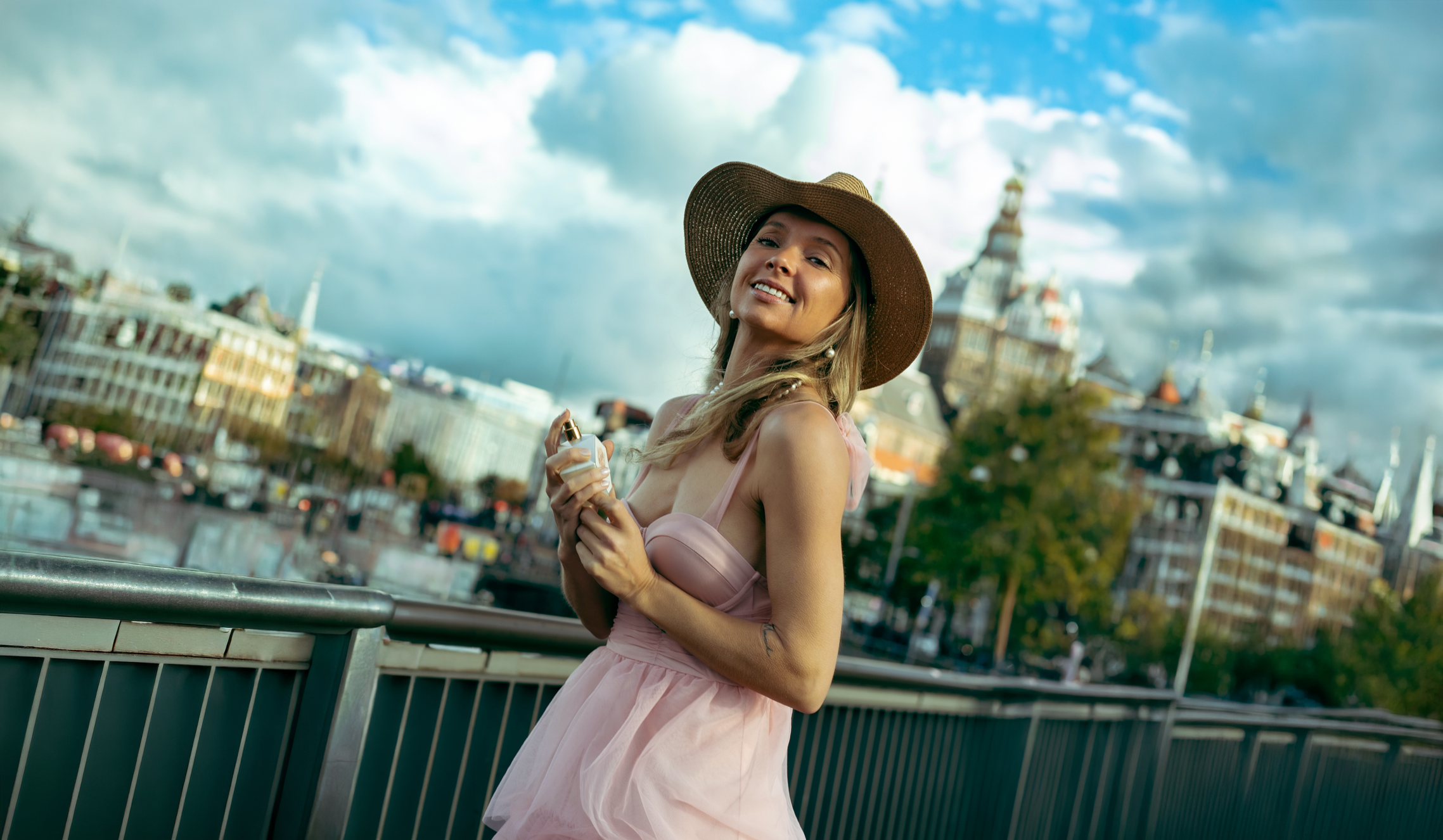 Woman in pink dress and straw hat holding By Jeske Dream perfume bottle on a bridge in Amsterdam